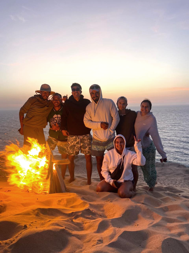 Groupe d’adultes autour d’un feu de camp sur la plage après un stage de surf, souriants avec Hatim